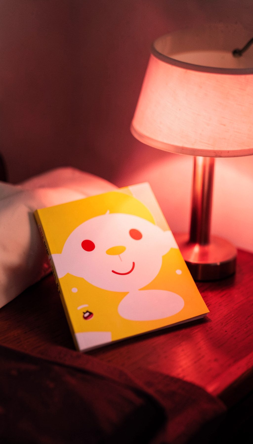 A trade paperback with a cheerful yellow and white cover, sitting on a nightstand next to a lamp, soft evening glow.