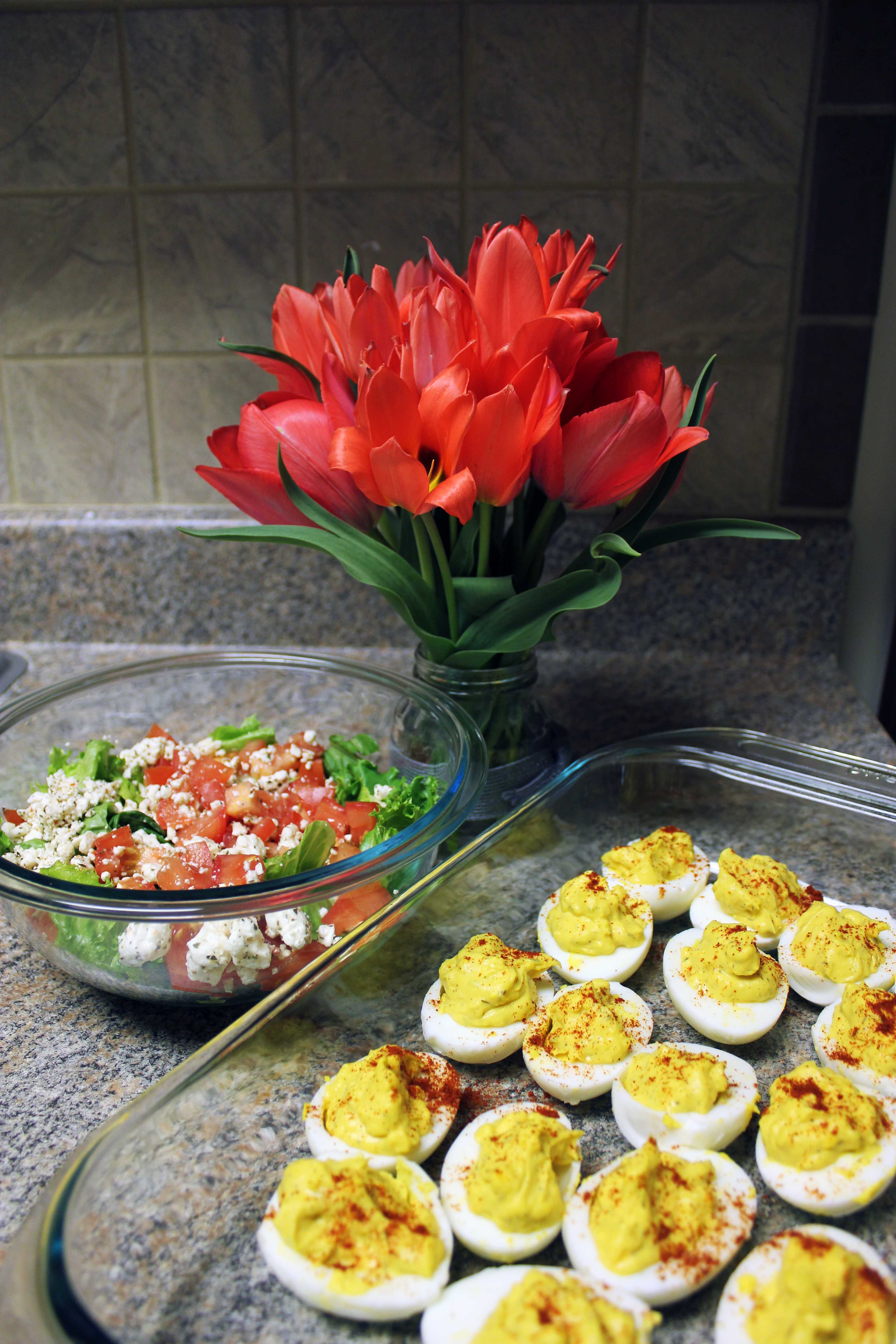 Red tulips in a vase beside a glass bowl of salad and a tray of deviled eggs on a granite counter, warm colorful kitchen