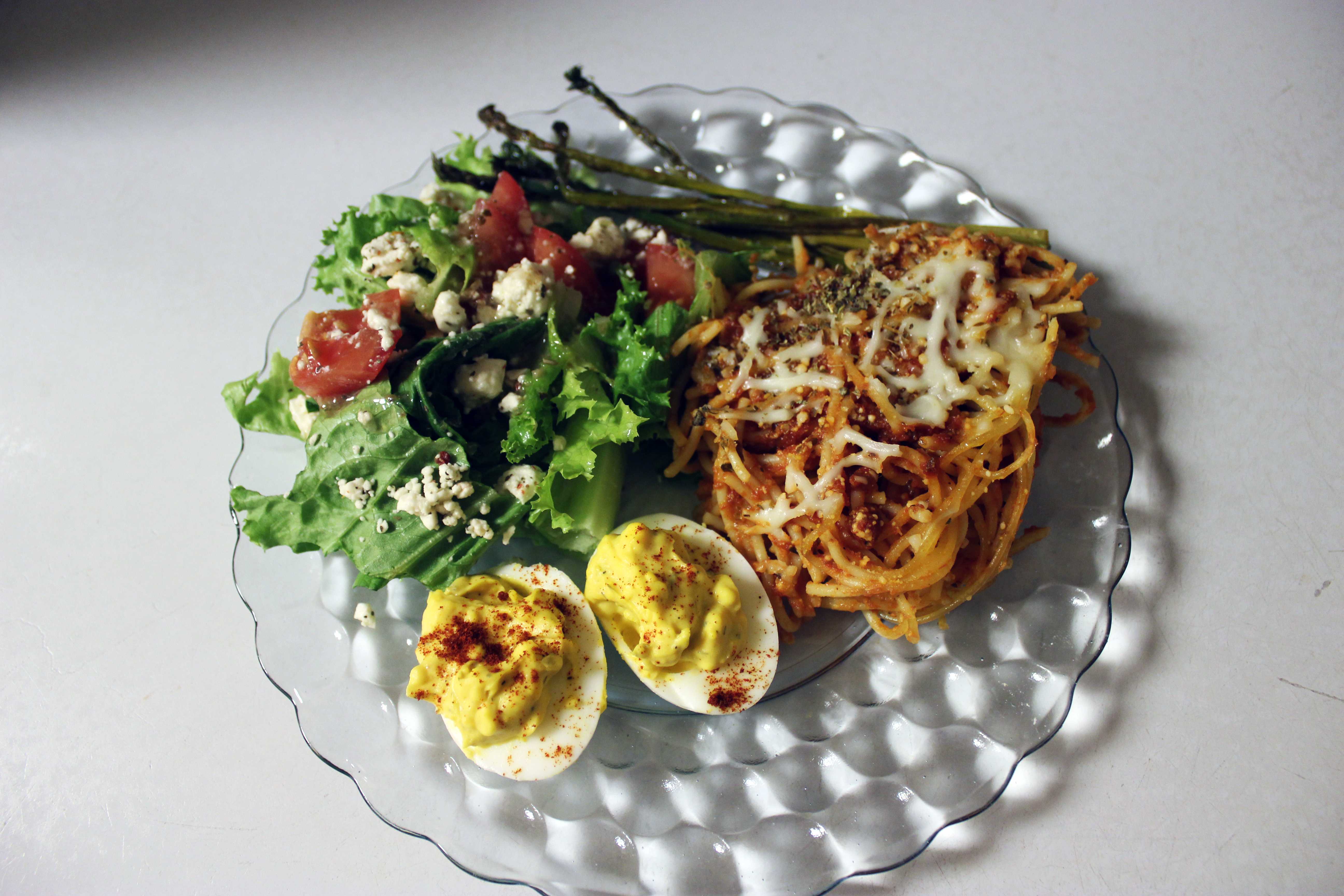 Plate of pasta with melted cheese, green salad with tomatoes and cheese crumbles, asparagus, and deviled egg halves