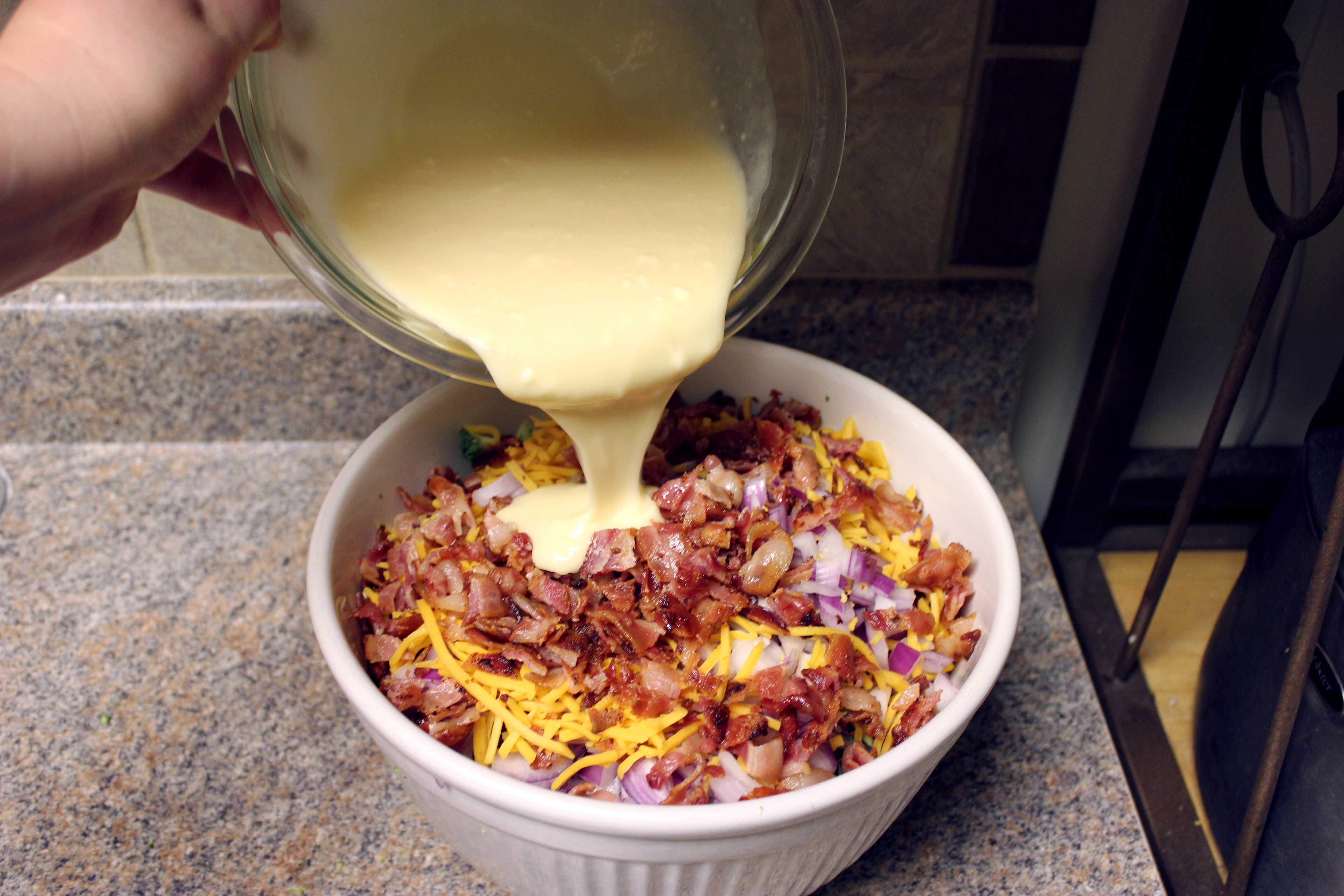 A creamy dressing being poured from a glass bowl into a large bowl filled with bacon bits, shredded cheese, and chopped broccoli