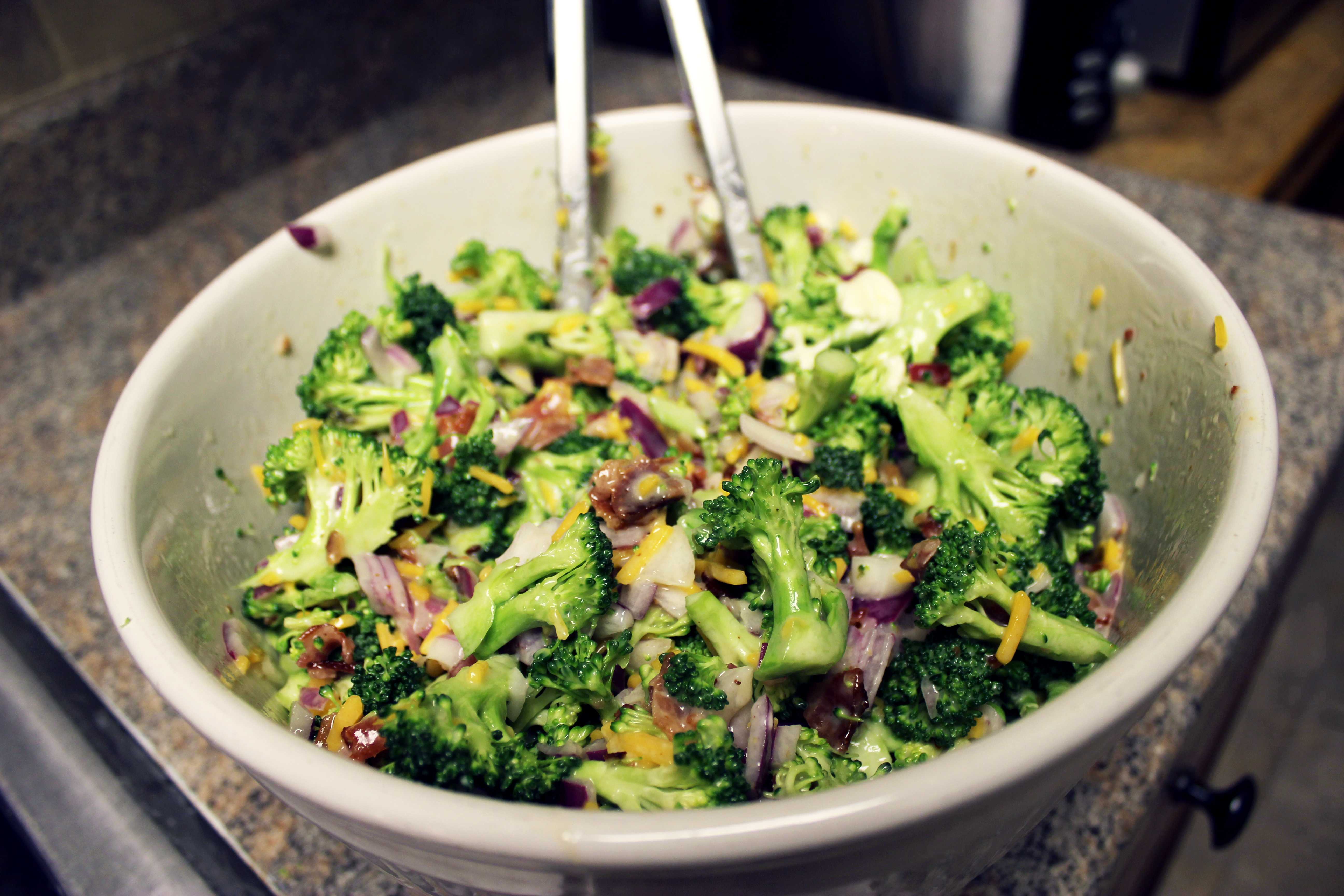 A large bowl of broccoli salad with red onion, cheese, and bacon bits, with serving tongs resting inside