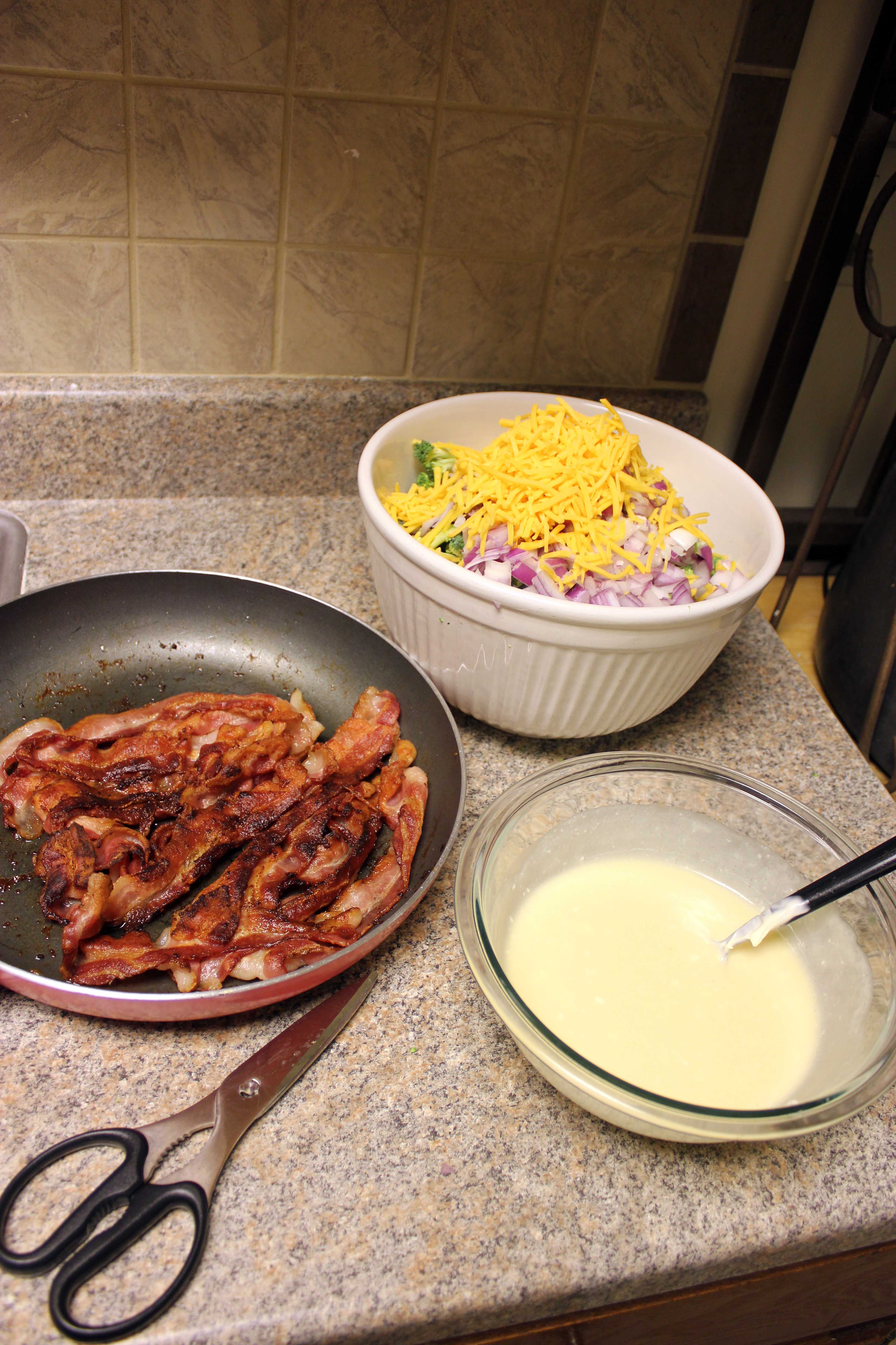 Kitchen counter with a pan of cooked bacon, a bowl of shredded cheese with chopped vegetables, and a bowl of creamy sauce