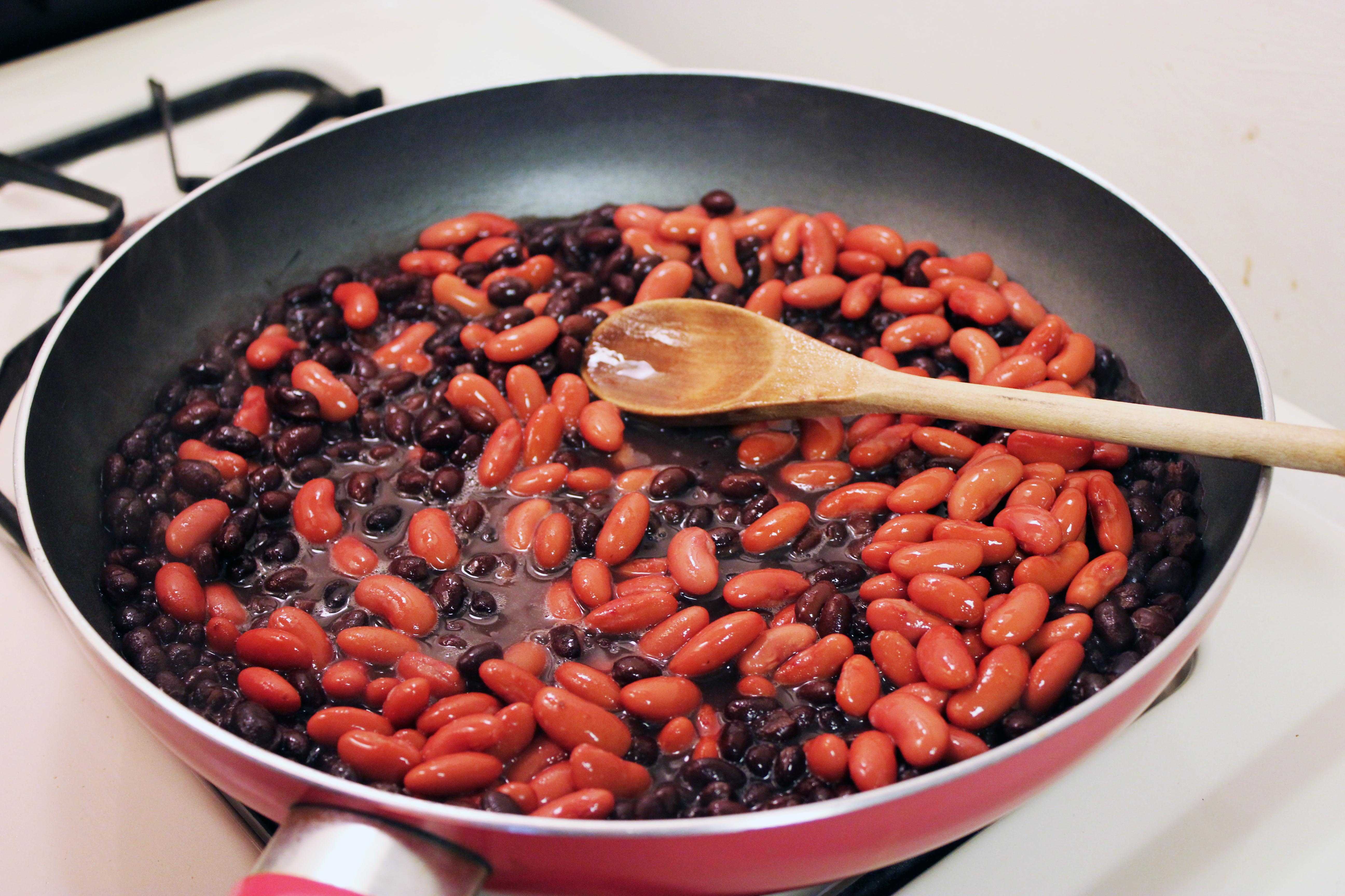 A skillet on a stovetop filled with cooked kidney beans and black beans with a wooden spoon