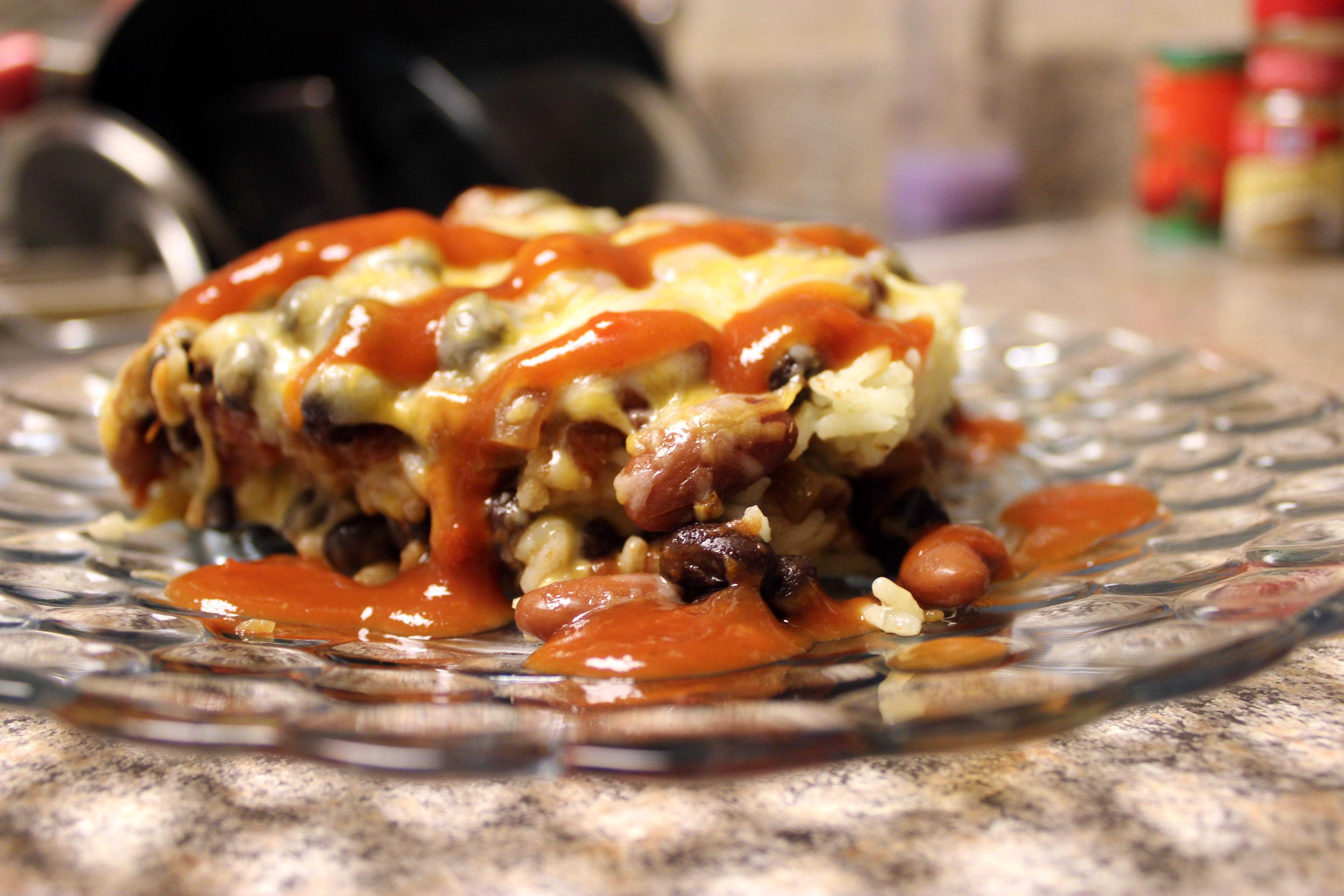 Close-up of a plated casserole slice with beans, rice, cheese and red sauce drizzled over it, on a glass plate