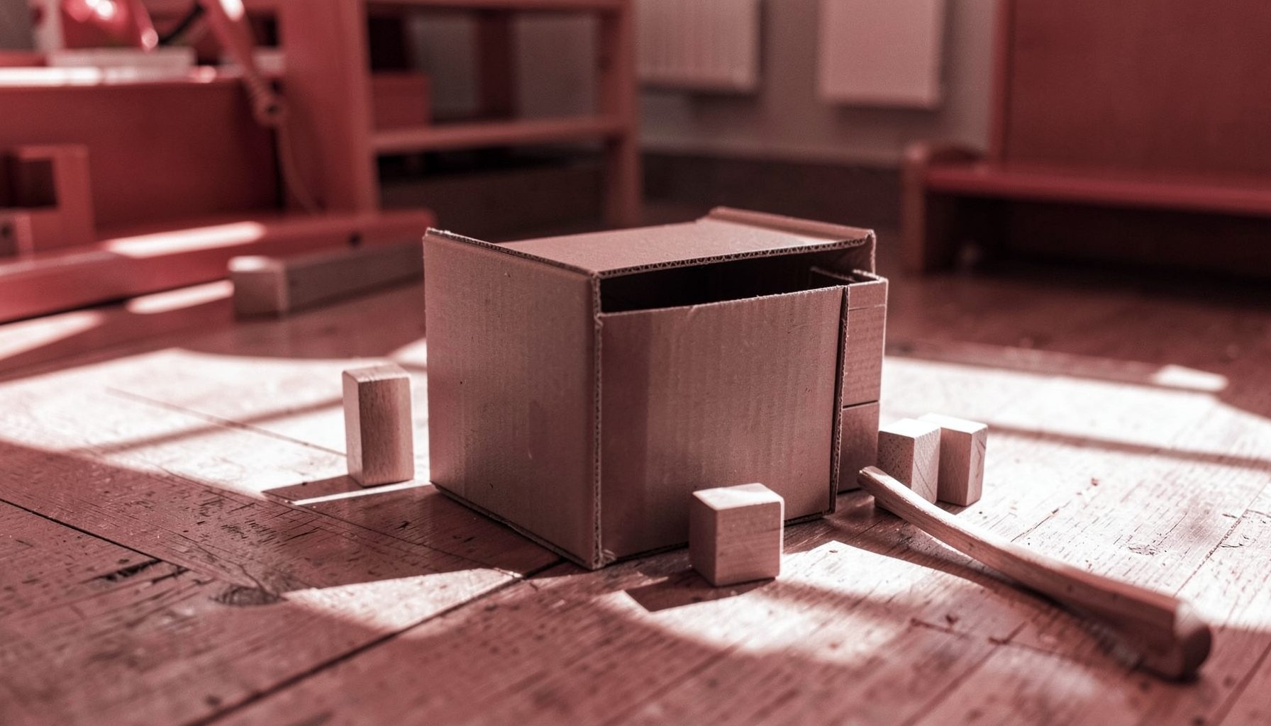 A cardboard box, wooden blocks, and a stick on a sunlit playroom floor, childhood simplicity
