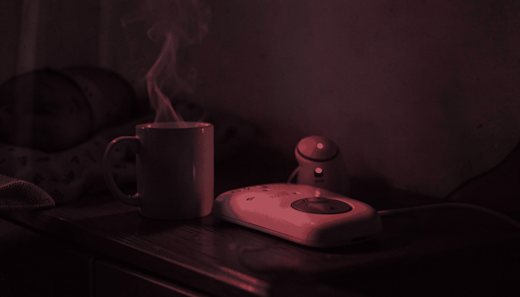 A steaming mug beside a baby monitor on a nightstand in dim early-morning light